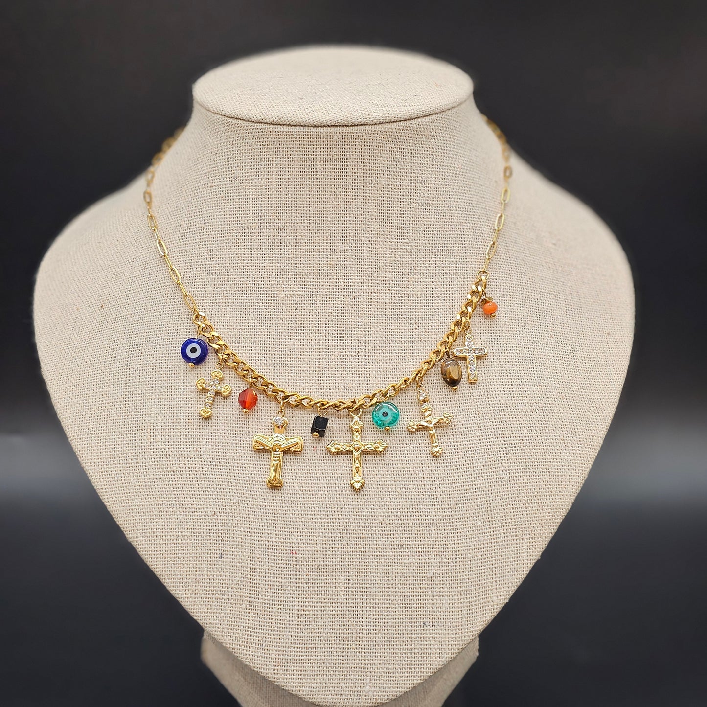 Vue d’ensemble du collier doré à charms croix et perles colorées sur présentoir beige.