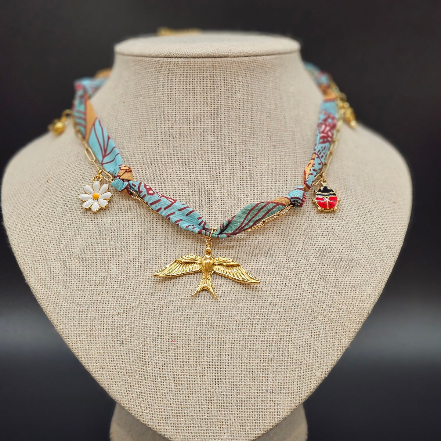 Collier foulard en tissu bleu et camel avec pendentif oiseau doré, marguerite et coccinelle, présenté sur buste beige.