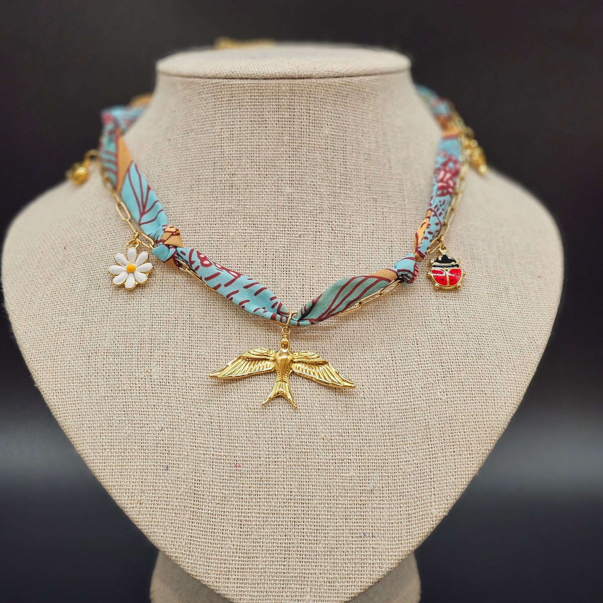 Collier foulard en tissu bleu et camel avec pendentif oiseau doré, marguerite et coccinelle, présenté sur buste beige.