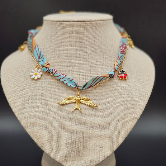 Collier foulard en tissu bleu et camel avec pendentif oiseau doré, marguerite et coccinelle, présenté sur buste beige.