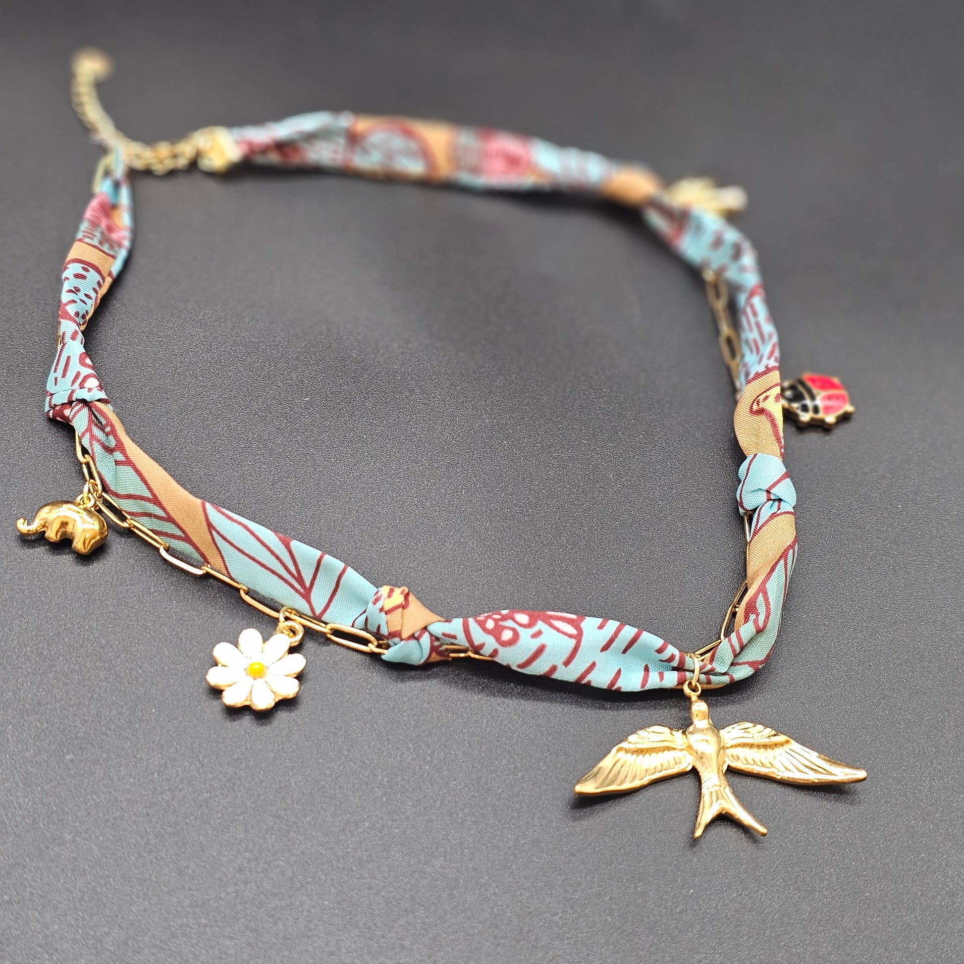 Collier foulard bleu et camel posé à plat sur fond noir, avec pendentif oiseau doré et charms marguerite, coccinelle et éléphant.